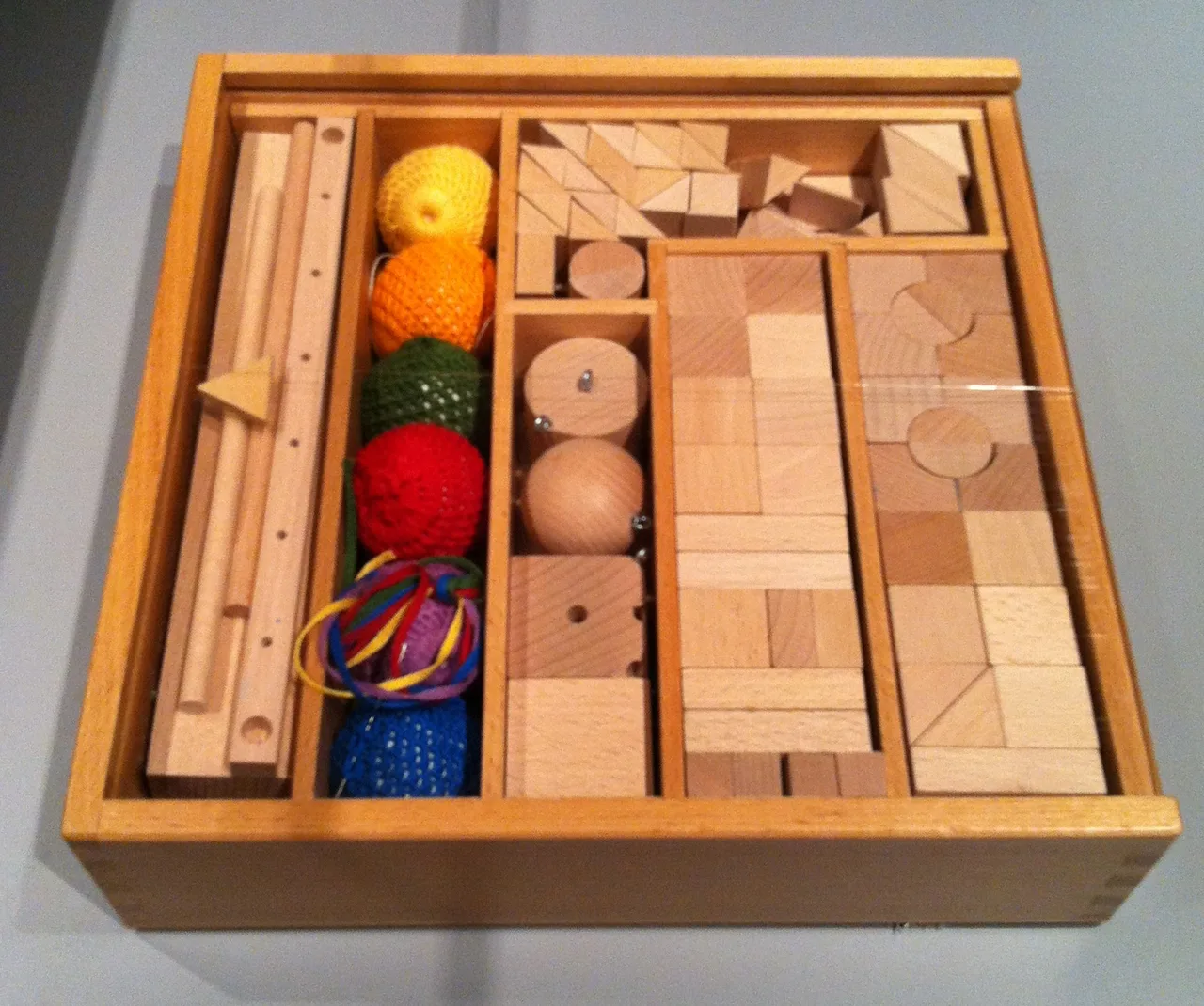 Caja de madera abierta con piezas geométricas de madera clara: cubos, prismas y tablillas ordenados en compartimentos, sobre fondo oscuro