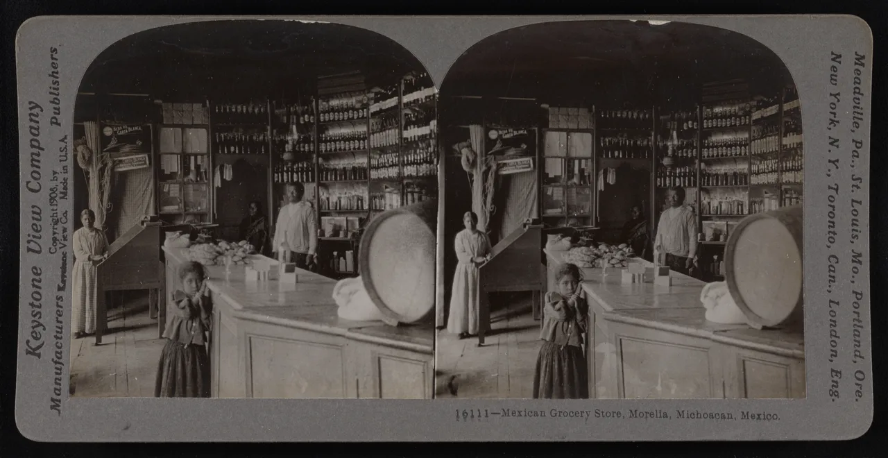 Una tienda de abarrotes, hacia 1908 Fotografía en blanco y negro de una tienda de abarrotes mexicana con estantes de madera llenos de mercancía y varios empleados detrás del mostrador