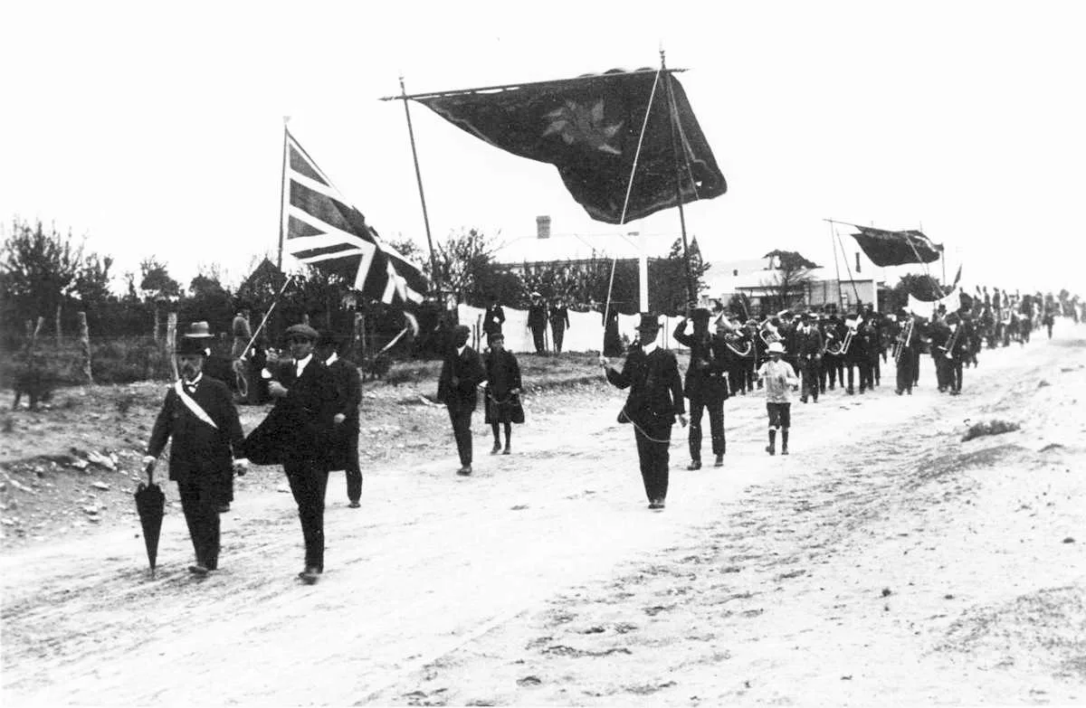 Fotografía antigua en sepia de una procesión: hombres con sombrero marchando por una calle de tierra detrás de grandes estandartes de tela